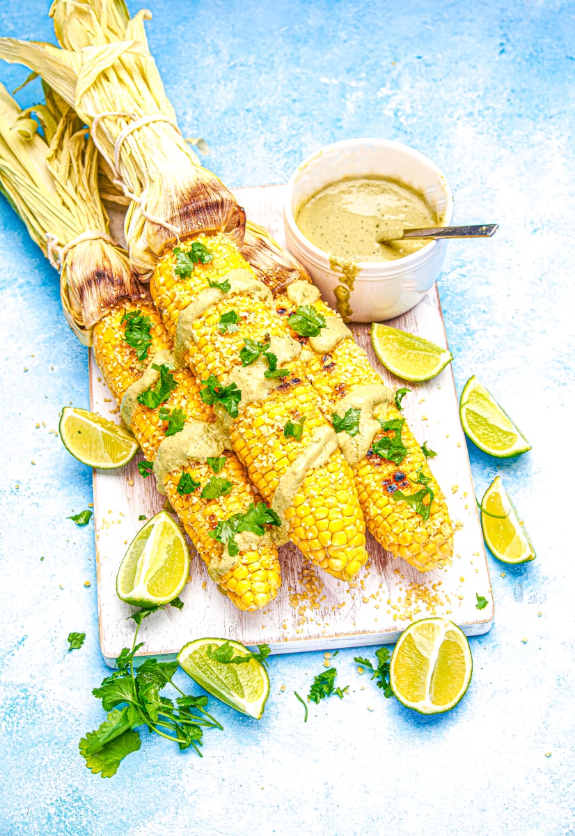 overhead view of Vegan Mexican Street Corn grilled in the husk on a white cutting board & blue table. 